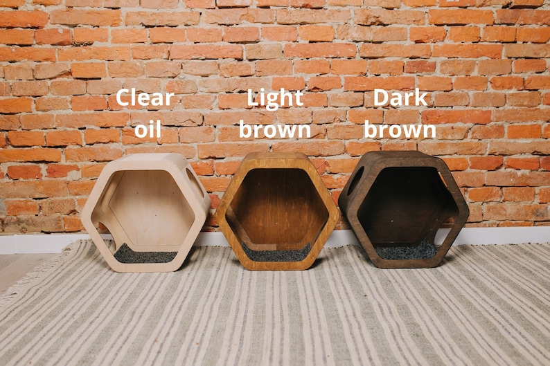 May include: Three hexagonal wooden pet beds in clear oil, light brown, and dark brown finishes. Each bed has a dark gray interior and is displayed against a brick wall. The text "Clear oil", "Light brown", and "Dark brown" is above each bed.