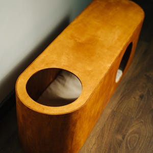 May include: A wooden pet bed with a warm, natural finish. The bed has an elongated, rounded shape with two circular openings. A light-colored cushion is visible inside. The bed sits on a wooden floor.