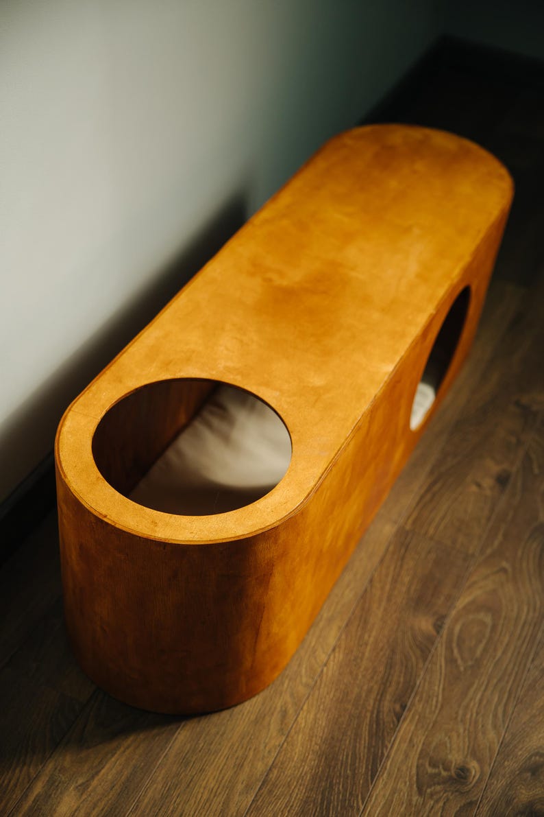 May include: A wooden pet bed with a warm, natural finish. The bed has an elongated, rounded shape with two circular openings. A light-colored cushion is visible inside. The bed sits on a wooden floor.