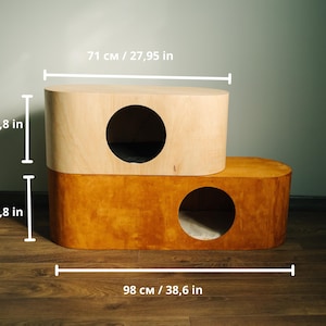 May include: A two-tiered cat house made of wood. The top level is light-colored, and the bottom level is a darker shade of wood. Each level has a circular opening. The cat house is 38.6 inches long, 27.95 inches wide, and 23.6 inches tall.