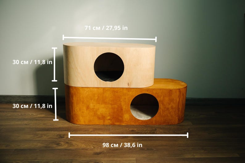 May include: A two-tiered cat house made of wood. The top level is light-colored, and the bottom level is a darker shade of wood. Each level has a circular opening. The cat house is 38.6 inches long, 27.95 inches wide, and 23.6 inches tall.
