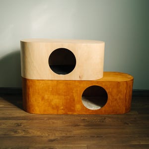 May include: A two-tiered cat house with a modern design. The top section is light-colored wood, and the bottom is a darker, orange-toned wood. Each level has a circular opening for entry.