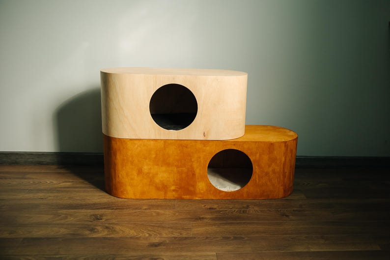May include: A two-tiered cat house with a modern design. The top section is light-colored wood, and the bottom is a darker, orange-toned wood. Each level has a circular opening for entry.