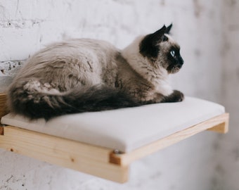 May include: A Siamese cat with blue eyes rests on a wall-mounted cat shelf. The shelf is made of light-colored wood and has a white cushion. The cat has a light grey body with dark brown markings and a long tail.