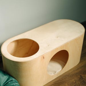 May include: A light-colored wooden cat house with two circular openings. One opening is on the top, and the other is on the side. The cat house has a rounded, oblong shape and sits on a wooden floor. A green beanbag is in the foreground.