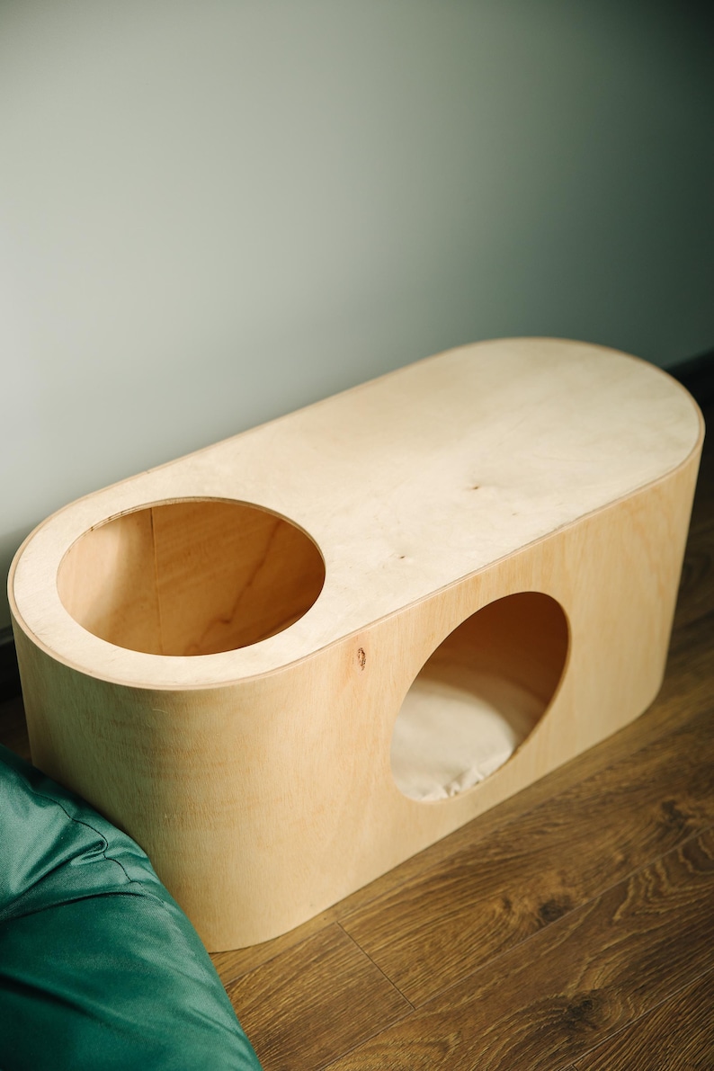 May include: A light-colored wooden cat house with two circular openings. One opening is on the top, and the other is on the side. The cat house has a rounded, oblong shape and sits on a wooden floor. A green beanbag is in the foreground.