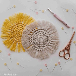 Pode incluir: Dois tapetes de crochê, um amarelo e outro bege, com franjas ao redor das bordas. Os tapetes estão sobre uma superfície branca com alfinetes e um gancho de crochê.