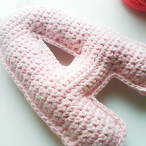 Puede incluir: Una letra A de crochet rosa. La letra está hecha con punto bajo y tiene una superficie ligeramente texturizada.
