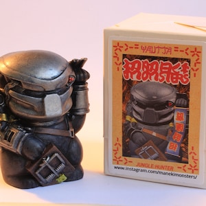 May include: A silver and black Predator action figure with a trap in its hand. The figure is standing on a white surface in front of a white box with Japanese text and the words "Jungle Hunter" and "www.instagram.com/manekimonsters/" printed on it.