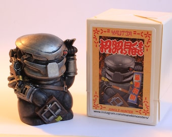 May include: A silver and black Predator action figure holding a trap. The figure is standing on a white surface in front of a white box with Japanese text and the words "Jungle Hunter" and "www.instagram.com/manekimonsters/" printed on it.
