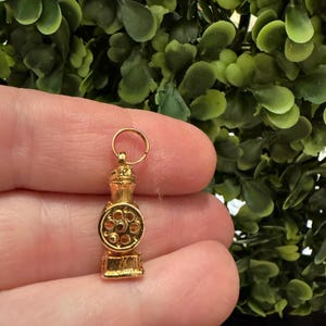 May include: A small, gold-colored metal charm shaped like a vintage coffee grinder. The charm features a cylindrical body with a circular front panel displaying a detailed gear or grinding mechanism and a small loop at the top for attachment.
