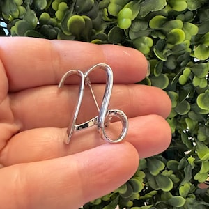 May include: A silver brooch in the shape of the Capricorn zodiac symbol. The brooch is held in a hand against a backdrop of green foliage. The design features the curved lines of the Capricorn symbol.