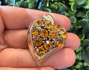 May include: A heart-shaped brooch with a silver tone finish and a cluster of amber-colored rhinestones.