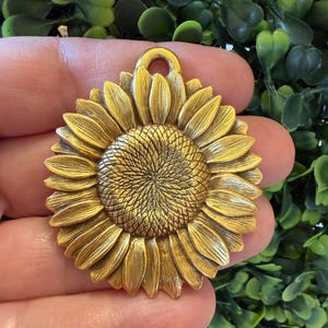 Könnte beinhalten: Ein kreisförmiger goldfarbener Metallanhänger mit einem detaillierten, geprägten Sonnenblumenmotiv, bei dem strukturierte Blütenblätter von einem zentralen Blütenkopf ausgehen. Der Anhänger hat eine kleine Öse zur Befestigung.