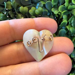 Peut inclure: Un pendentif en forme de cœur avec une finition nacrée. Le pendentif présente un motif en fil de couleur or en forme de la lettre "V" et des volutes décoratives. Le cœur est tenu dans une main sur un fond de feuillage vert.