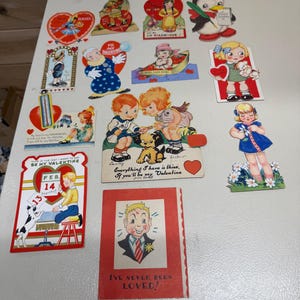 May include: A collection of vintage Valentine's Day cards and decorations. The cards feature illustrations of children, animals, and hearts. Some cards have text, including "Teacher," "Be My Valentine," and "I've Never Been Loved!"