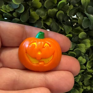May include: A small, orange, jack-o'-lantern pumpkin charm with a bright green stem. The pumpkin features a carved, smiling face with yellow cutouts for the eyes, nose, and mouth, creating a cheerful expression suitable for seasonal decoration.