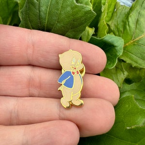 Op de afbeelding: Een gouden metalen pin met een cartoon varken dat een blauw jasje en een rode strik draagt. Het varken heeft een roze neus en kijkt naar rechts.
