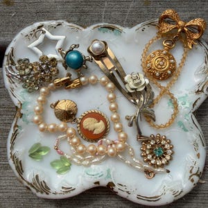 May include: A collection of assorted jewelry pieces including a pearl necklace, a cameo pendant, a gold-tone bow brooch, a turtle pin, a star charm, and floral-themed hair clips, all resting on a white scalloped decorative tray with gold trim.