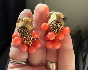 May include: A pair of gold-tone metal earrings featuring a textured, tulip-shaped top component. Multiple fine gold-tone chains hang from the base, each ending in a small, round, coral-colored bead, creating a clustered, dangling effect.
