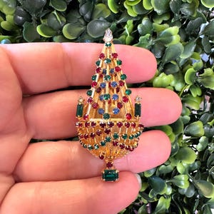 Op de afbeelding: Een goudkleurige kerstboom broche, versierd met rode, groene en blauwe edelstenen. De broche heeft een gedetailleerd ontwerp met een sprankelende heldere edelsteen aan de bovenkant en een groene rechthoekige edelsteen aan de basis. Groene bladeren op de achtergrond.