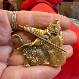 Könnte beinhalten: Goldfarbene Metallbrosche einer Katze, die Cello spielt. Die Katze trägt einen formellen Blazer und hält einen Bogen. Die Brosche hat aufwendige Details und eine Vintage-Ästhetik. Der Schwanz der Katze kringelt sich um ihren Körper.