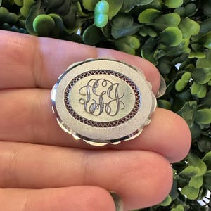 Puede incluir: Broche ovalada de plata con un monograma grabado. La broche tiene un borde festoneado y un diseño detallado alrededor del monograma central. El monograma está en un estilo de escritura elegante.