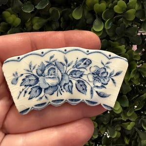 May include: A white porcelain decorative piece with a blue floral design. The piece has a scalloped edge and a detailed pattern of roses and other flowers. The blue and white color scheme gives it a classic, vintage look.