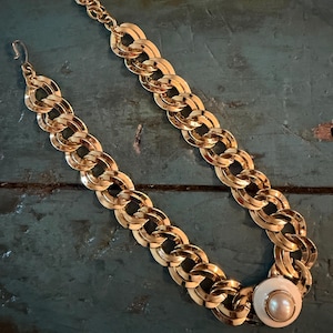 May include: A gold-tone chain link necklace featuring a series of interlocking oval links. The necklace includes an adjustable chain extension with a hook clasp and a circular pendant at one end, which contains a central white pearl-like bead.