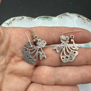 May include: A pair of silver dangle earrings featuring a delicate, cutout design of a woman in a flowing dress. The earrings have a simple hook closure.