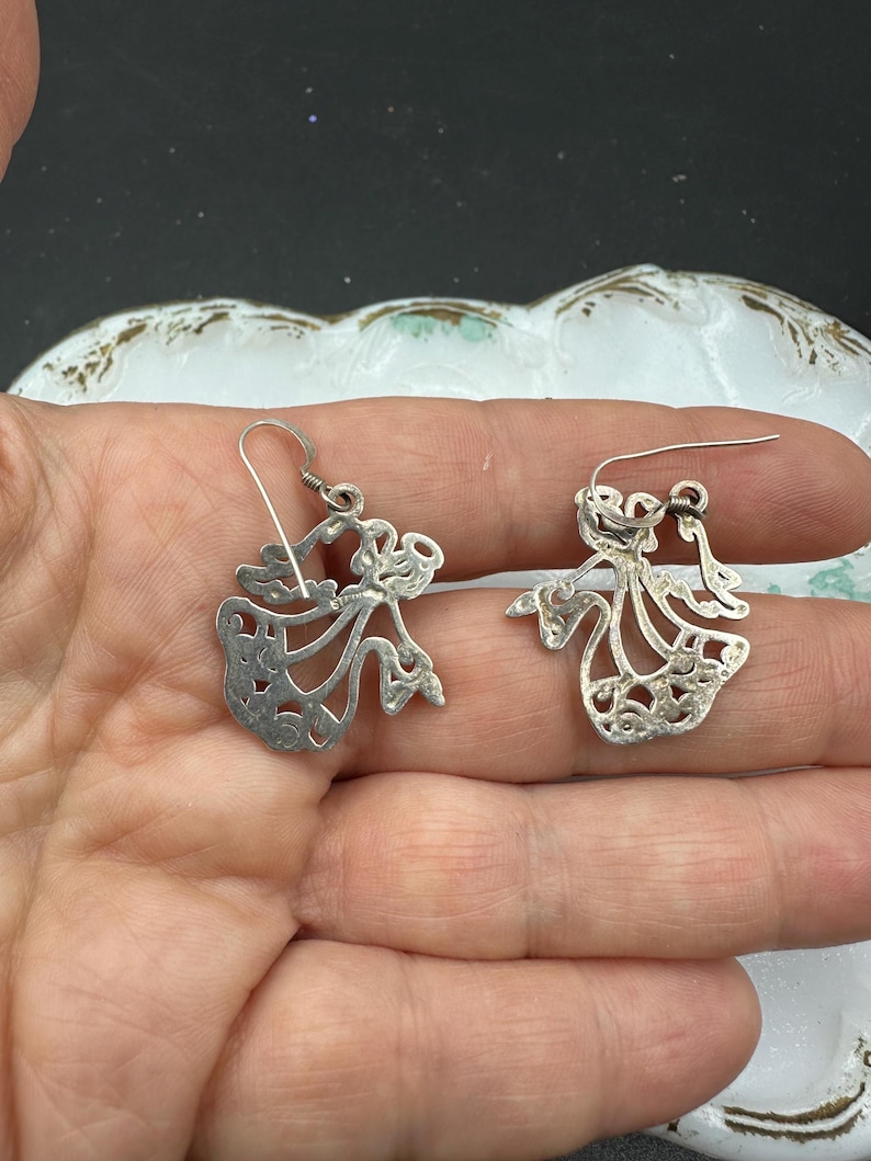 May include: A pair of silver dangle earrings featuring a delicate, cutout design of a woman in a flowing dress. The earrings have a simple hook closure.
