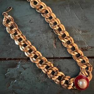 May include: A gold-tone chain link necklace featuring a series of interlocking oval links. One end of the necklace terminates in a circular pendant with a red enamel border surrounding a central white faux pearl. A hook clasp is at the other end.