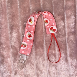 May include: A pink and white fabric dummy clip with a strawberry pattern and a clear plastic clip. The clip has a red ribbon attached to it.