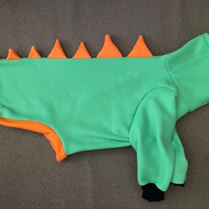 Peut inclure: Un pull pour animal de compagnie vert vif avec une rang&eacute;e de pointes triangulaires orange le long du dos. Le v&ecirc;tement poss&egrave;de des manches longues avec des poignets noirs, un col haut et un liser&eacute; orange sur le bas, rappelant un costume de dinosaure.