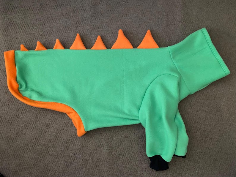 Peut inclure: Un pull pour animal de compagnie vert vif avec une rang&eacute;e de pointes triangulaires orange le long du dos. Le v&ecirc;tement poss&egrave;de des manches longues avec des poignets noirs, un col haut et un liser&eacute; orange sur le bas, rappelant un costume de dinosaure.