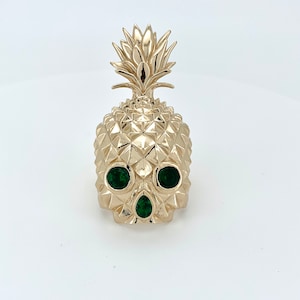 May include: A gold pineapple skull ring with green gemstone eyes.