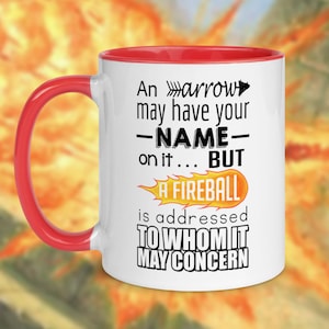 Puede incluir: Taza de café de cerámica blanca con borde rojo. La taza tiene un gráfico negro con el texto "An arrow may have your -NAME- on it... BUT A FIREBALL is addressed TO WHOM IT MAY CONCERN".