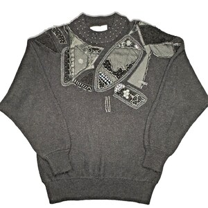 May include: A dark grey sweater with decorative patches on the shoulders and chest. The patches feature sequins and beads. The sweater has a crew neck and long sleeves. The measurements are in centimetres.