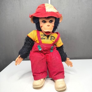 May include: A plush monkey doll with black fur, wearing a red and cream bucket hat, a yellow t-shirt with the text ZIP printed in black, and red corduroy overalls featuring green buttons. The doll has light-coloured plastic hands and shoes.