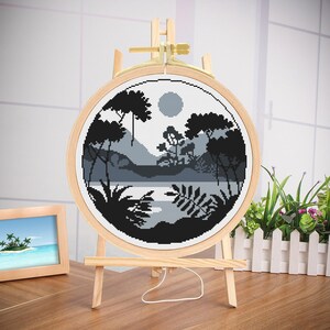 May include: A black and white cross-stitch embroidery of a forest scene with a lake and a full moon. The embroidery is framed in a round wooden hoop and displayed on a wooden easel.