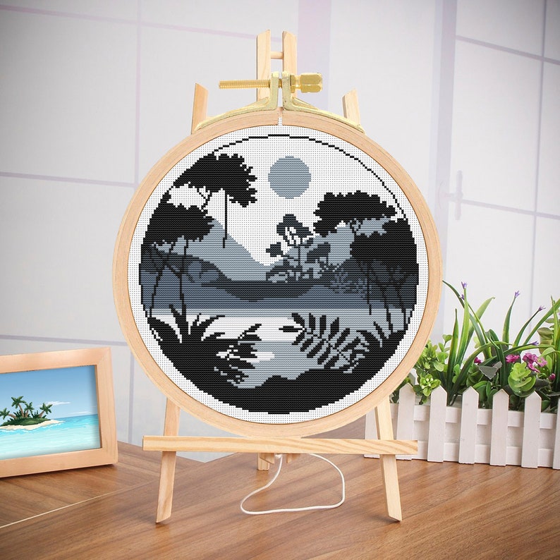 May include: A black and white cross-stitch embroidery of a forest scene with a lake and a full moon. The embroidery is framed in a round wooden hoop and displayed on a wooden easel.