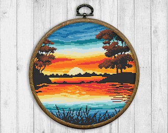 May include: A round wooden embroidery hoop featuring a cross-stitch design of a sunset over a lake with two trees on the shore. The sky is blue and orange, and the water is blue and green.