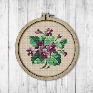 May include: A cross-stitch embroidery of a bouquet of purple violets with green leaves in a wooden embroidery hoop.