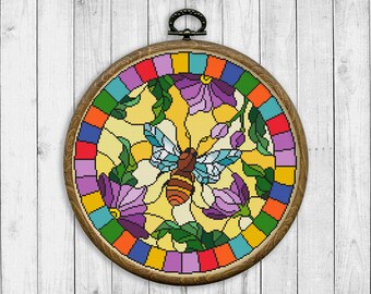 Könnte beinhalten: Ein runder Stickrahmen mit einem farbenfrohen Kreuzstich-Design einer Biene, die zwischen lila Blumen fliegt. Das Design erinnert an Buntglas.