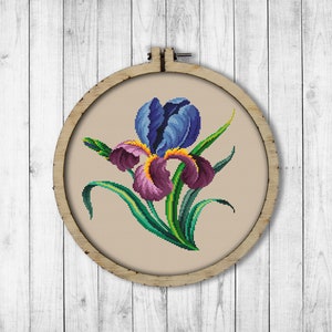 May include: A cross-stitch embroidery of a purple and blue iris flower with green leaves in a wooden embroidery hoop.