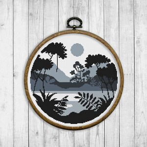 May include: A black and white cross-stitch embroidery of a landscape scene with trees, mountains, and a lake. The embroidery is framed in a round wooden hoop.