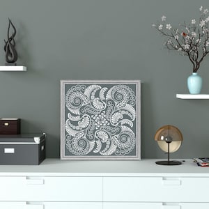 Op de afbeelding: Een wit ingelijste canvas print met een grijze achtergrond en een wit bloemenontwerp. De print hangt aan een witte muur boven een witte commode met drie laden. Er staat een klein zwart doosje op de commode, een zwarte lamp met een gouden bol aan de rechterkant van de commode en een klein wit plankje met een blauwe vaas en een tak met witte bloemen aan de linkerkant van de commode.