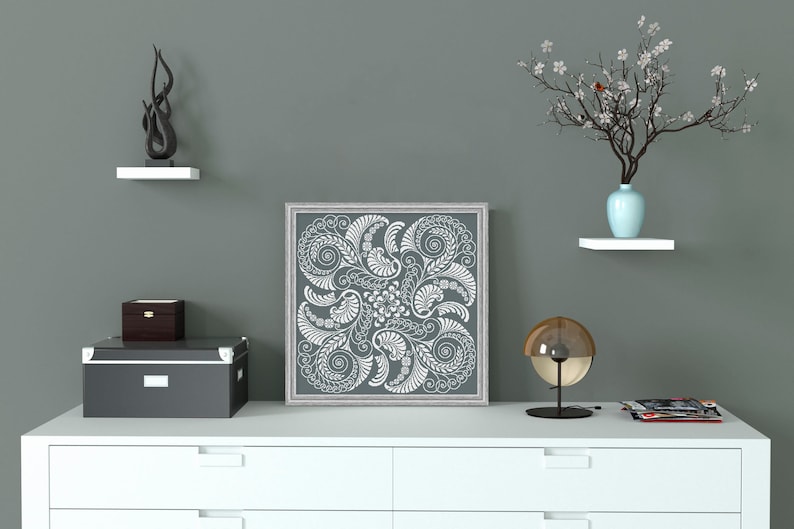 Op de afbeelding: Een wit ingelijste canvas print met een grijze achtergrond en een wit bloemenontwerp. De print hangt aan een witte muur boven een witte commode met drie laden. Er staat een klein zwart doosje op de commode, een zwarte lamp met een gouden bol aan de rechterkant van de commode en een klein wit plankje met een blauwe vaas en een tak met witte bloemen aan de linkerkant van de commode.