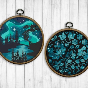 May include: Two round embroidery hoops with dark wood frames. The left hoop features a cross-stitch design of a forest scene with a river and a starry sky with a blue and green aurora borealis. The right hoop features a cross-stitch design of a floral pattern with blue and green flowers on a black background.