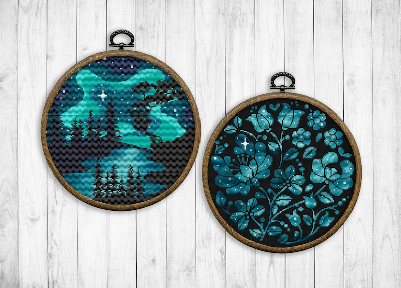 May include: Two round embroidery hoops with dark wood frames. The left hoop features a cross-stitch design of a forest scene with a river and a starry sky with a blue and green aurora borealis. The right hoop features a cross-stitch design of a floral pattern with blue and green flowers on a black background.